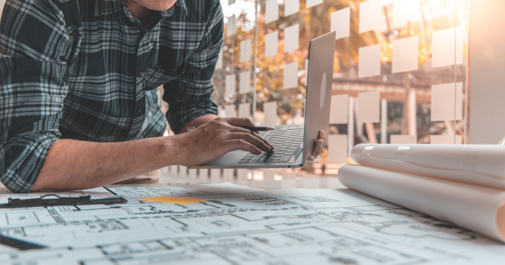 Architect working on a laptop with architectural blueprints and rolled-up plans on a desk, showcasing modern engineering and construction planning.