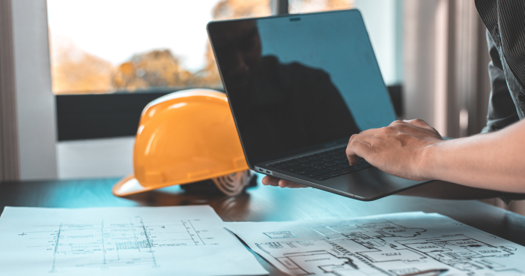 Engineer reviewing digital building plans on a laptop with printed blueprints and a safety helmet on the desk, representing modern engineering and construction planning.